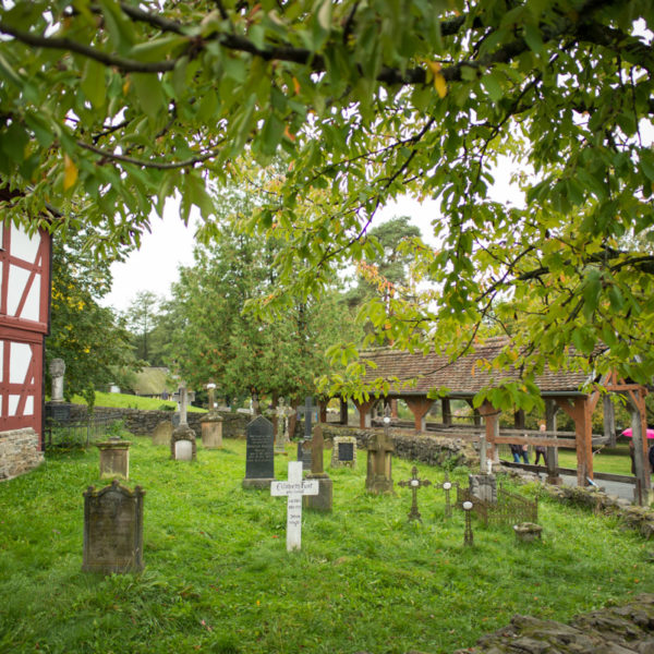 Blick auf den Kirchhof der Kirche aus Niederhörlen mit gesammelten Grabsteinen. Im VOdergrund ein weißes Kreuz.