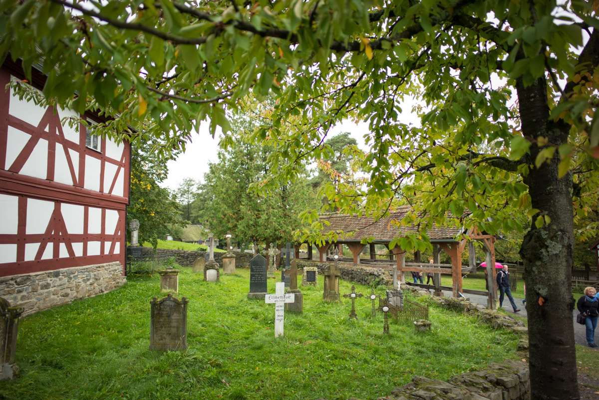Blick auf den Kirchhof der Kirche aus Niederhörlen mit gesammelten Grabsteinen. Im VOdergrund ein weißes Kreuz.