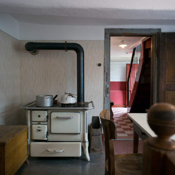 Stube im Erdgeschoss mit Blick auf die Tür, danaben ein alter Holzkochherd. An der linken Wand steht eine Truhe, darüber hängt eine kleine Gaderobe.