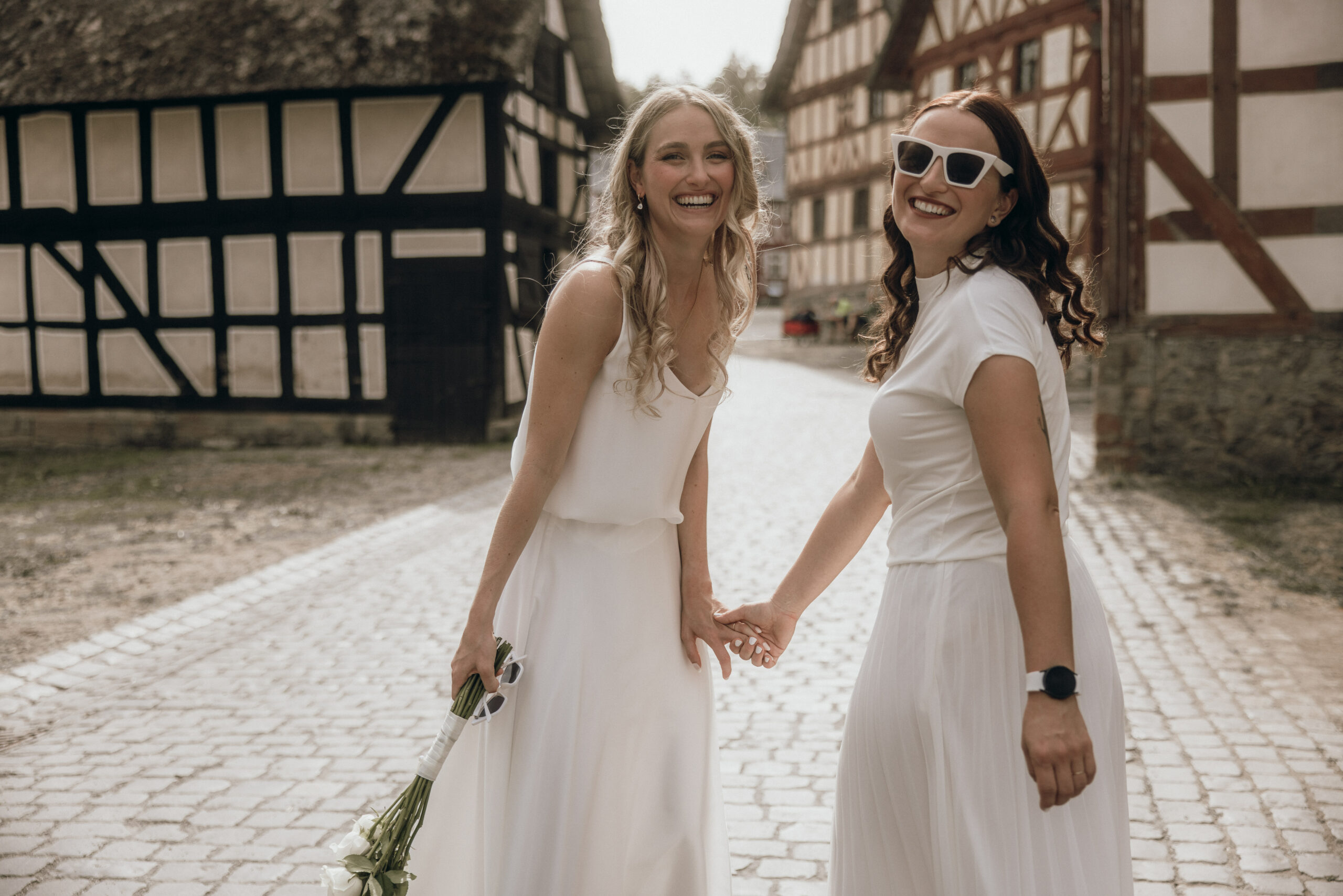 Glückliches Hochzeitspaar: zwei Frauen schauen in die Kamera und stehen vor Fachwerkhäusern.