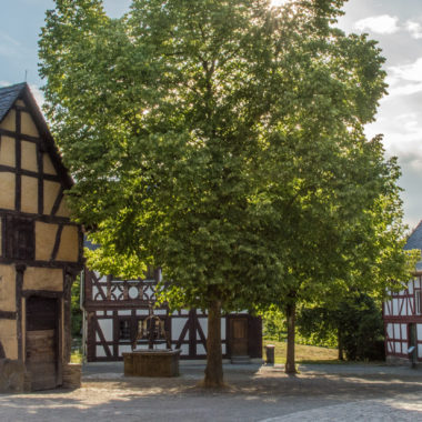 Dorfplatz in der Baugruppe Mittelhessen, man sieht einen Lindenbaum, die alte Schmiede aus Weinbach und die Kirche aus Niederhörlen.