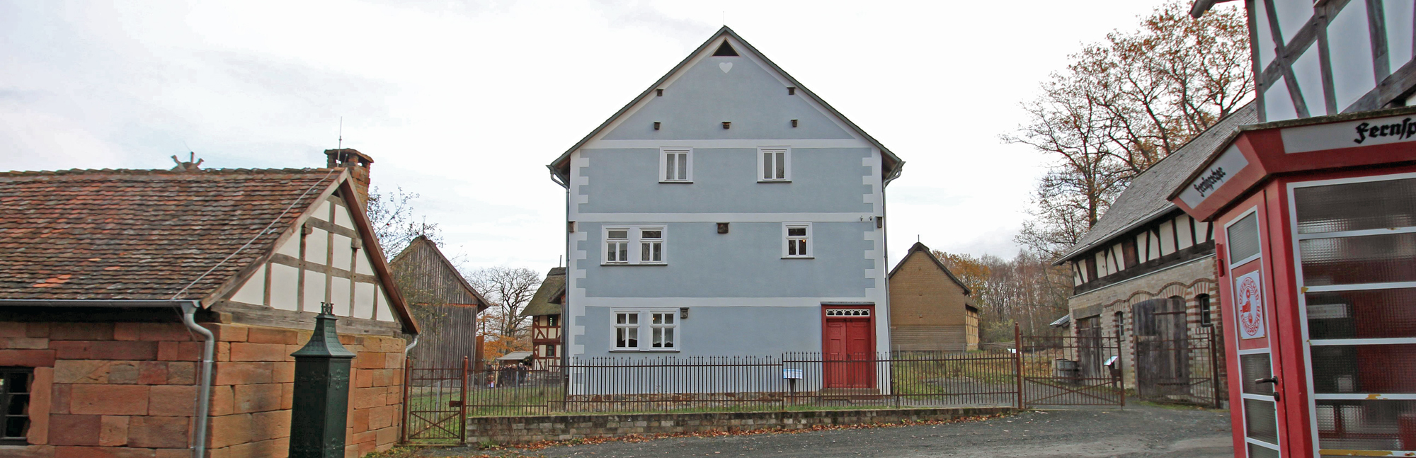 Außenansicht Haus aus Breitenbach, dreistöckig mit hellblauem Anstrich und roter Eingangstür. Davor ein Metallstabzaun, auf der rechten Seite ist die Telefonzelle vor dem Posthaus zu erkennen.