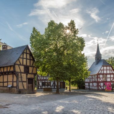 Dorfplatz im Sommer. Man sieht die Schmiede aus Weinbach, die Kirche aus Niederhörlen und die Linde mit dichtem Blätterdach.