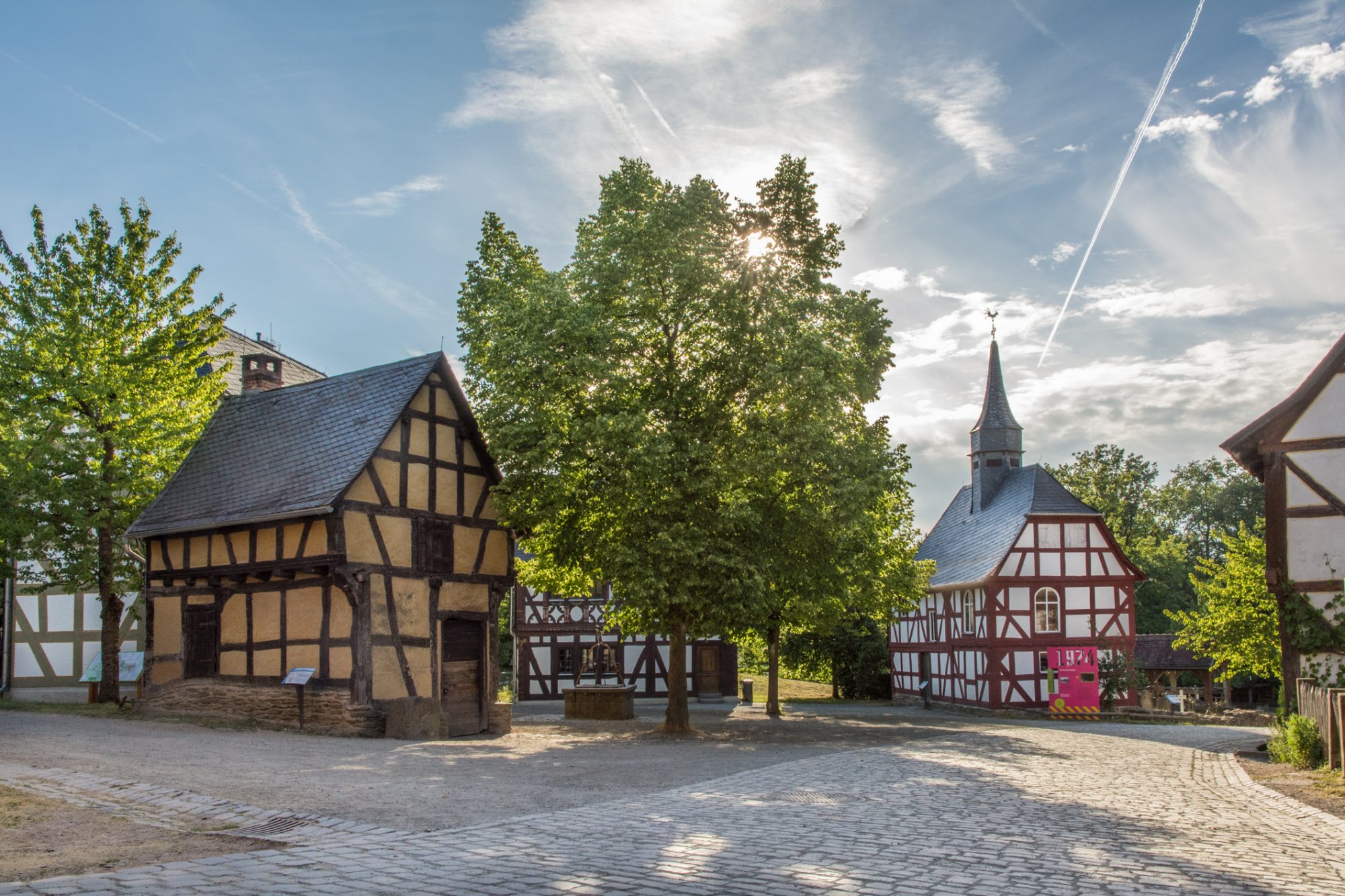 Dorfplatz im Sommer. Man sieht die Schmiede aus Weinbach, die Kirche aus Niederhörlen und die Linde mit dichtem Blätterdach.