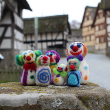 Gefilzte Clowns stehen auf einem Stein, im Hintergrund ist die Dorfstraße zu sehen.