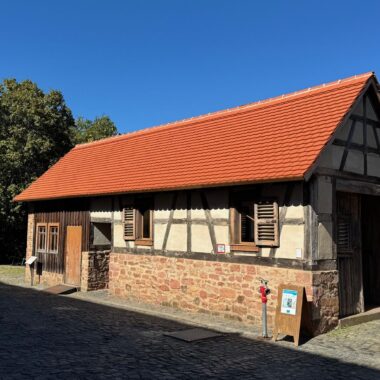 Historisches Fachwerkhaus mit rotem Ziegeldach auf Kopfsteinpflaster. Das Gebäude zeigt traditionelle deutsche Bauweise mit dunklen Holzbalken, heller Füllung und Fensterläden. Links große Bäume, rechts weiteres historisches Gebäude, im Vordergrund eine Informationstafel.