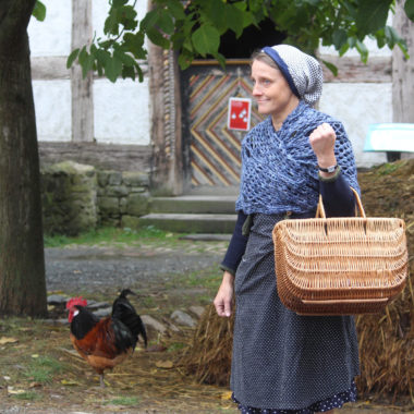 Eine Schauspielerin des Museumstheaters schlüpft in die Rolle von Frieda Sommer, eine ehemalige Bewohnerin des Hauses aus Eisemroth. Sie steht vor einem Misthaufen. Im Hintergrund laufen Hühner und man sieht den Eingang zu einem historischen Gebäude.