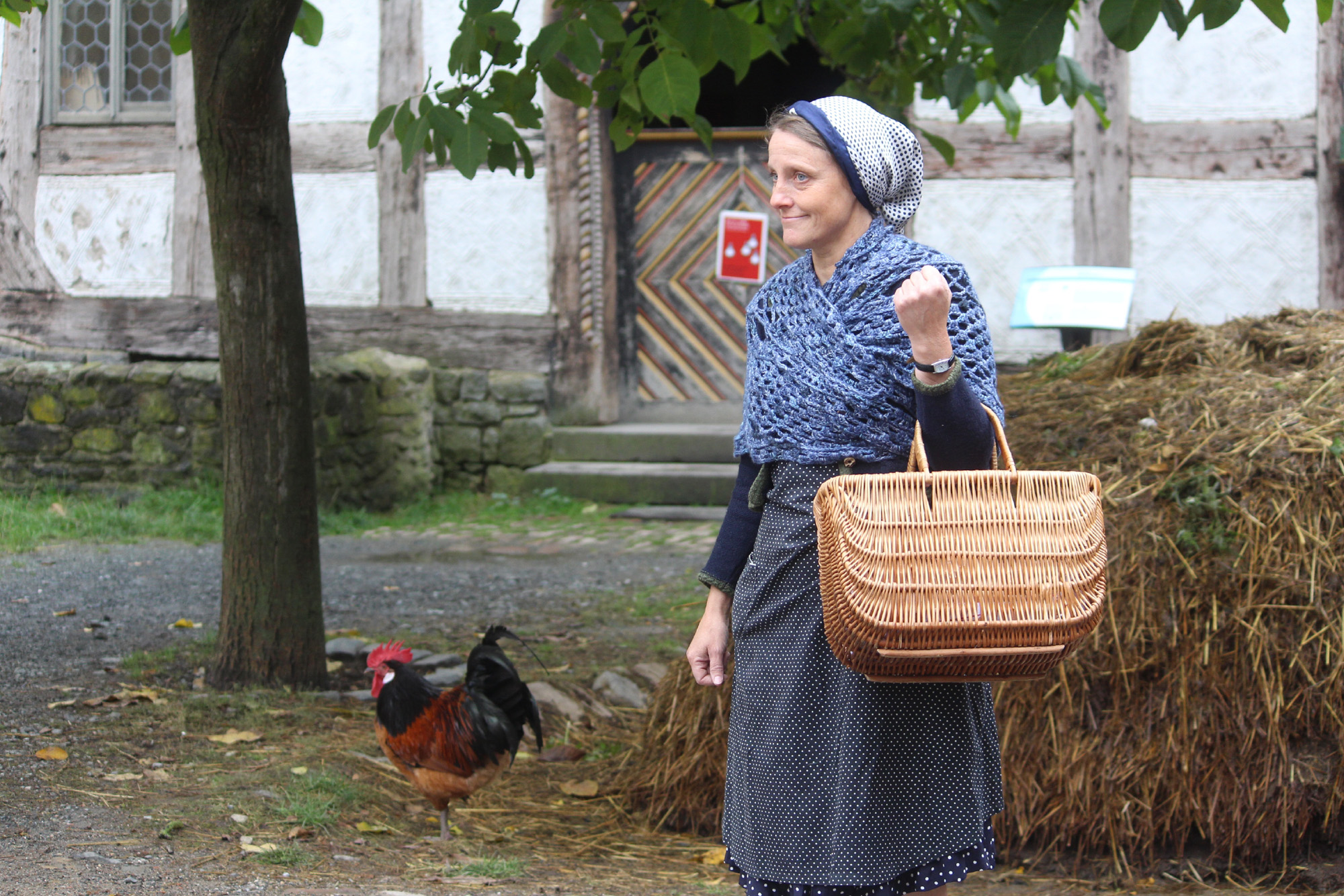 Eine Schauspielerin des Museumstheaters schlüpft in die Rolle von Frieda Sommer, eine ehemalige Bewohnerin des Hauses aus Eisemroth. Sie steht vor einem Misthaufen. Im Hintergrund laufen Hühner und man sieht den Eingang zu einem historischen Gebäude.