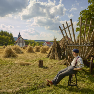 Getreideernte im Hessenpark. Ein Erntehelfer sitzt auf einem Schemel und ruht sich aus, im Hintergrund steht ein Erntewagen und verschiedene Getreidegarben.