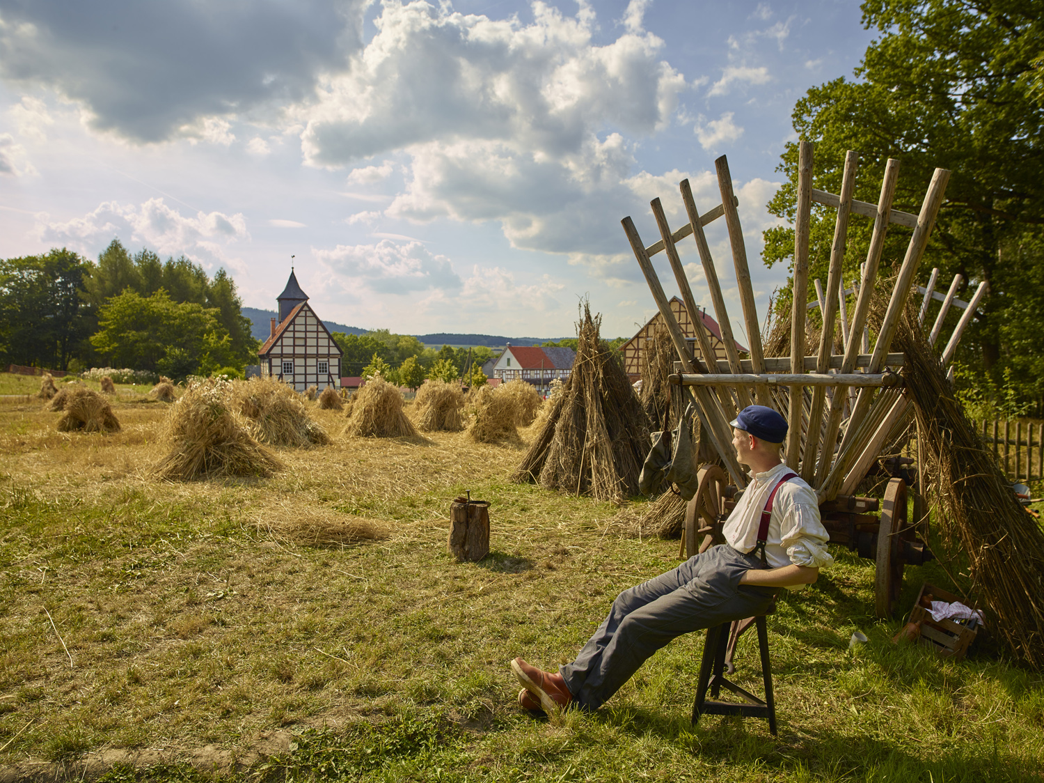 Getreideernte im Hessenpark. Ein Erntehelfer sitzt auf einem Schemel und ruht sich aus, im Hintergrund steht ein Erntewagen und verschiedene Getreidegarben.