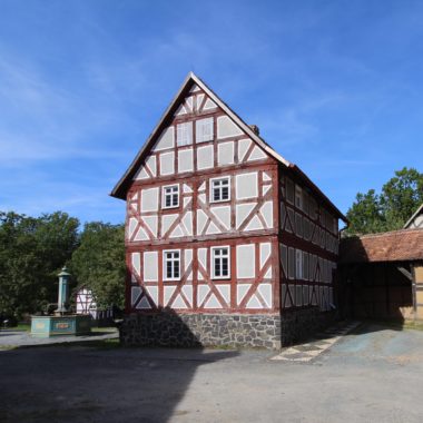 Ein rotes Fachwerkhaus mit zwei Stockwerken. Links daneben ein Brunnen.