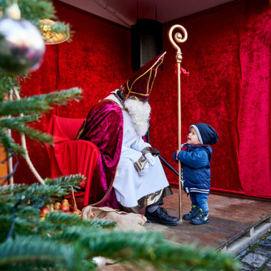 Sankt Nikolaus sitzt auf seinem Stuhl und blickt zu einem kleinen Jungen herab.