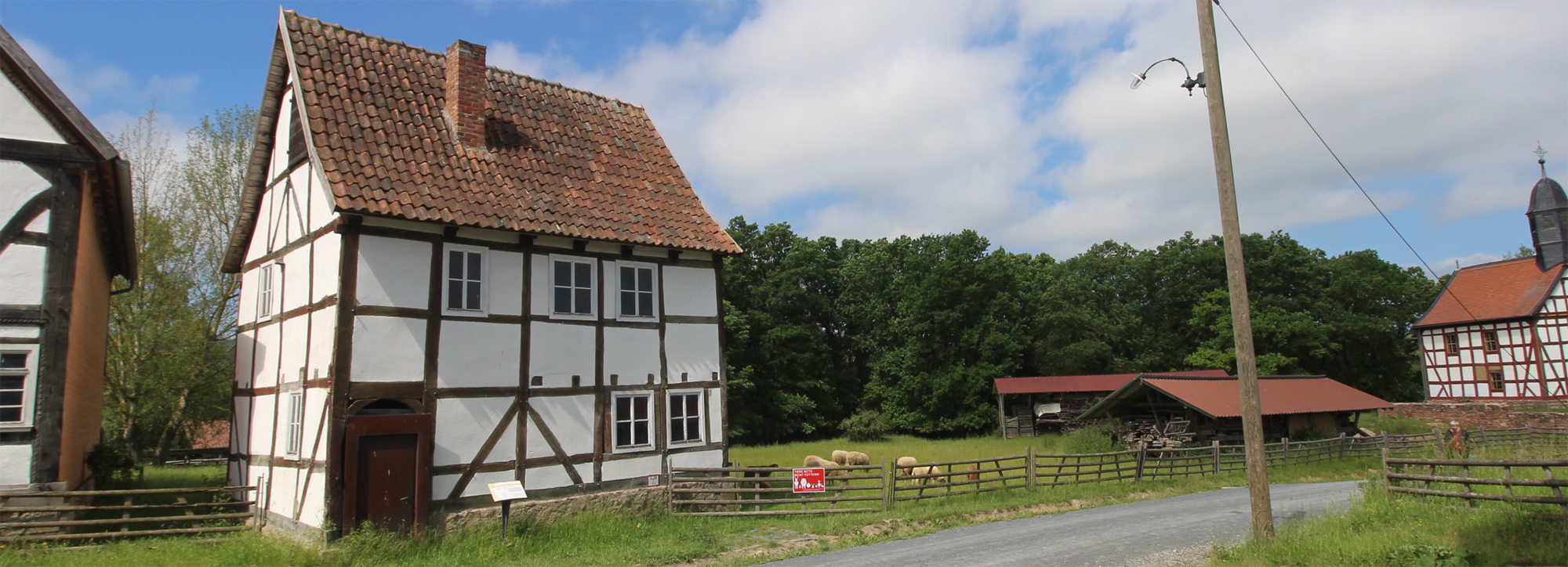 Ein kleines Fachwerkhaus mit Ziegeldach, rechts daneben eine große Wiese mit Schafen.