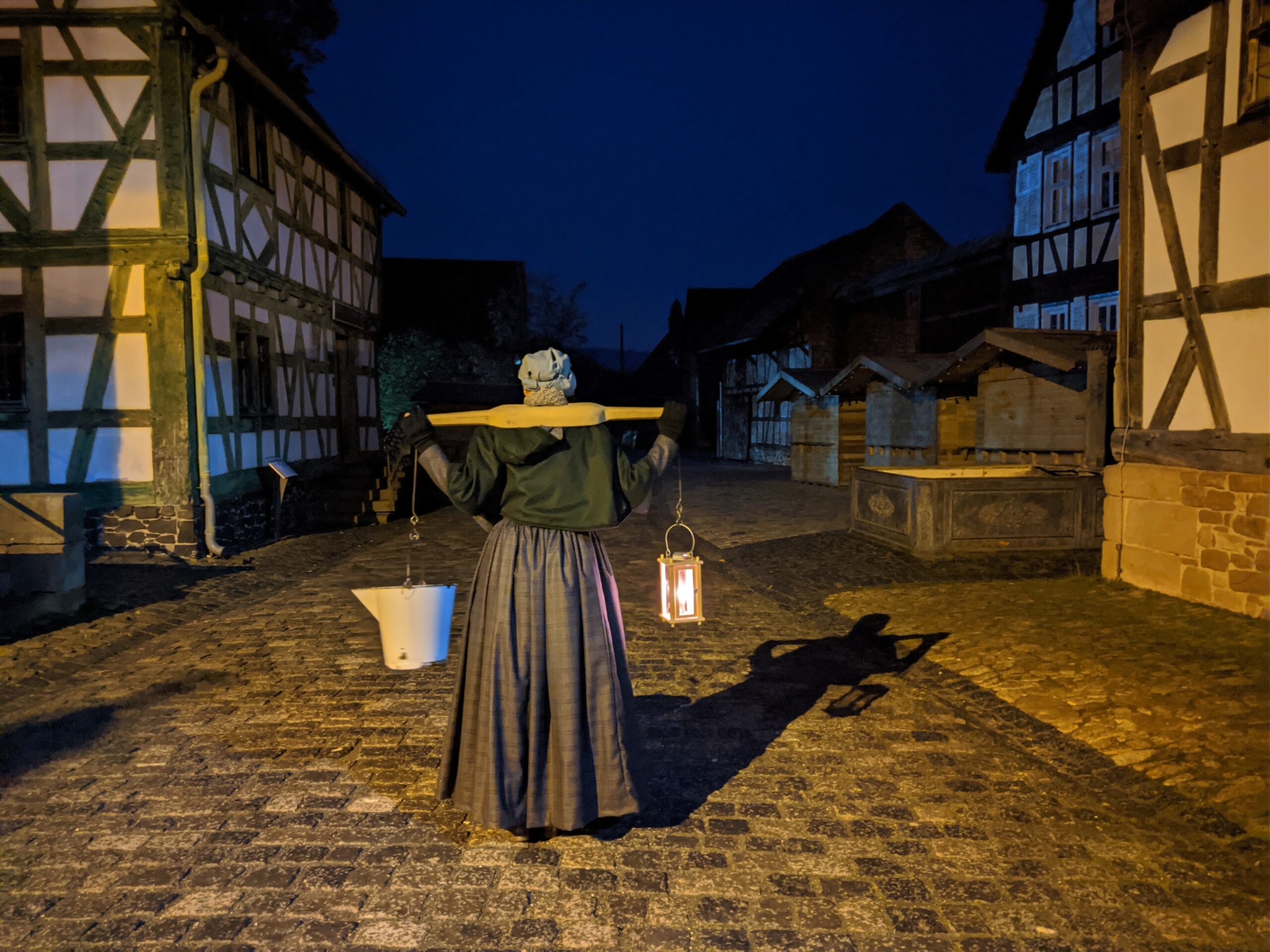 Schauspielerin läuft in der Dunkelheit die Dorfstraße entlang. Auf den Schultern trägt sie ein Wasserjoch. Man sieht sie von hinten.