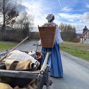 Frau im historischen Kostüm, voll bepackt mit Kiepe und Handwagen.