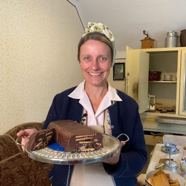 Historische Hausfrau hat eine Kuchenplatte in der Hand, darauf die Kuchenspezialität 