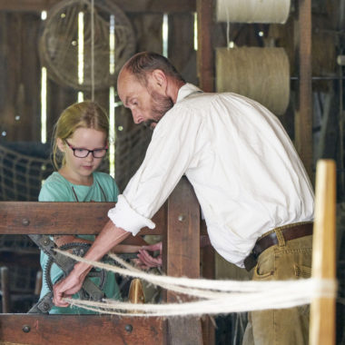 Der Vorführhandwerker stellt gemeinsam mit einem Mädchen ein Seil her. Das Mädchen kurbelt und der Seiler führt die einzelnen Stränge zu einem Seil zusammen.