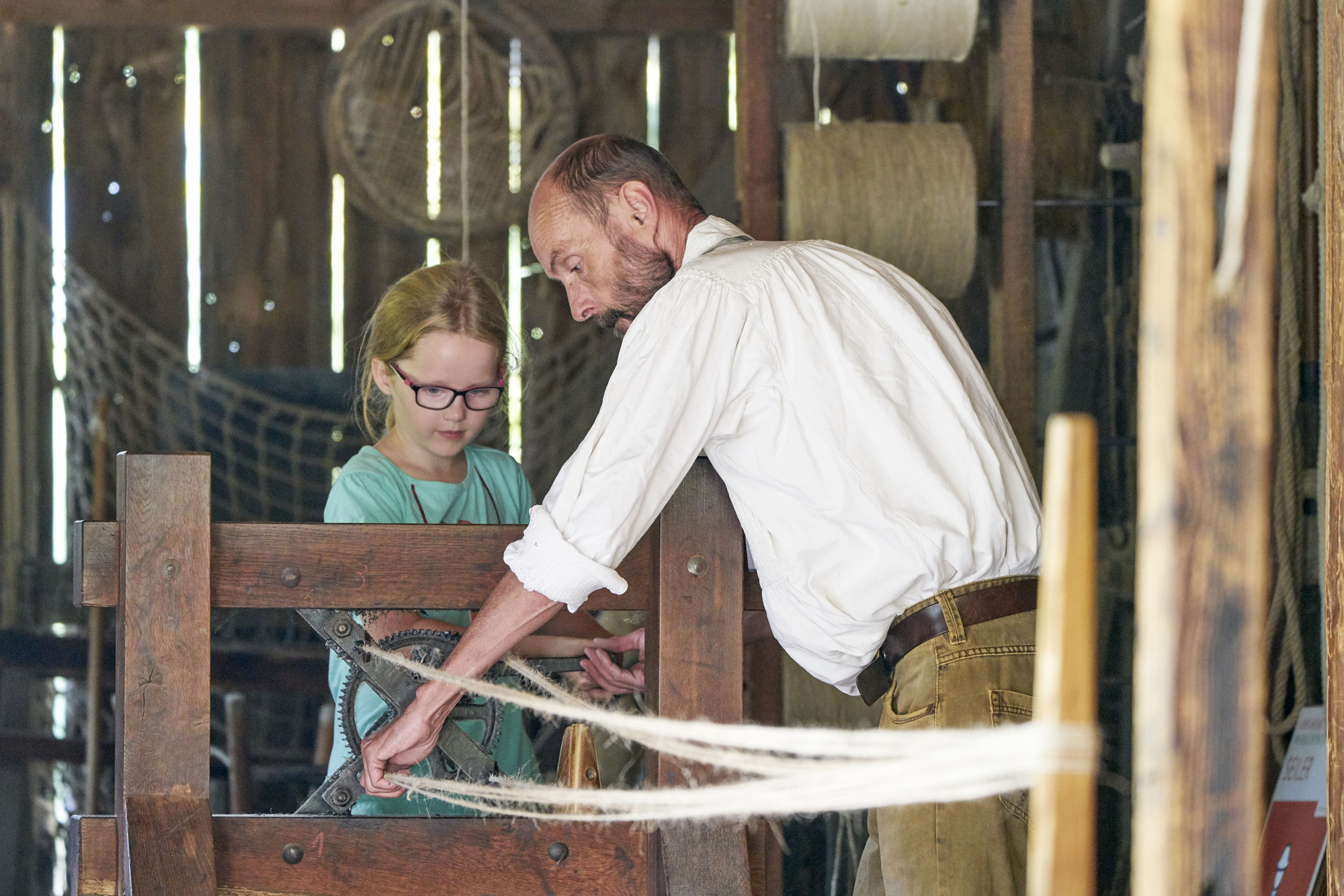 Der Vorführhandwerker stellt gemeinsam mit einem Mädchen ein Seil her. Das Mädchen kurbelt und der Seiler führt die einzelnen Stränge zu einem Seil zusammen.