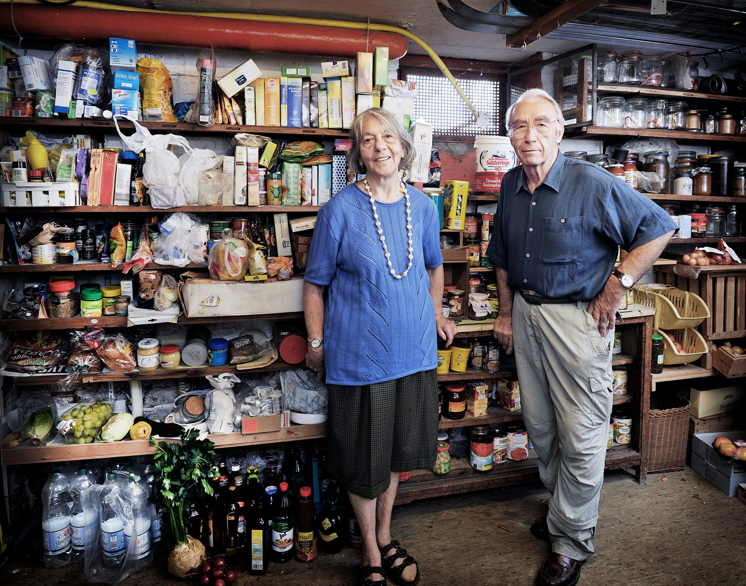 Zwei ältere Menschen stehen vor einem grossen Regal, in dem Nahrungsvorräte gelagert sind.