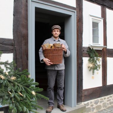 Bäcker mit Mütze hält einen Weidenkorb voller Brot in der Tür eines Fachwerkhauses