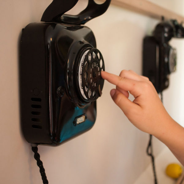 Telefon mit Wählscheibe an einer weißen Wand befestigt. Eine Person wählt an der Drehscheibe