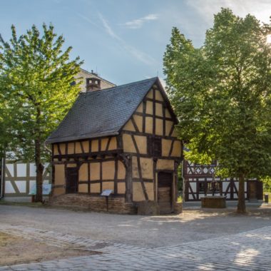 Die Schmiede aus Weinbach steht in der Baugruppe Mittelhessen am Dorfplatz. Sie wird umrahmt von zwei grünen Bäumen.