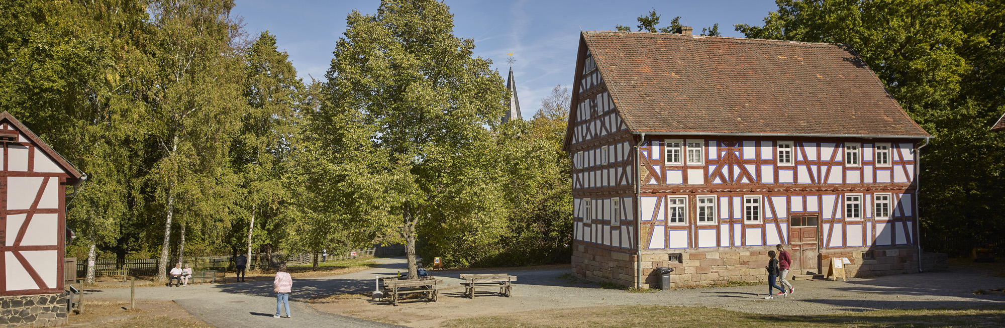 Fachwerkhaus aus Sterzhausen mit gemauertem Sockel. Links daneben ein großer Laubbaum, darunter Sitzbänke